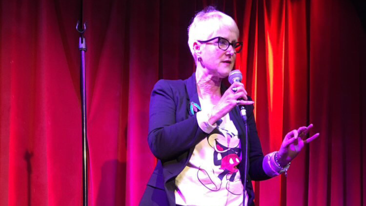 A woman with short hair and glasses speaks into a microphone with red stage curtains behind her.