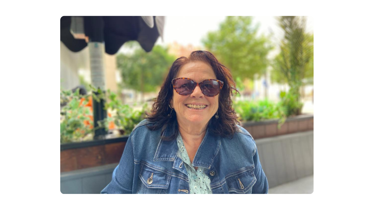 A woman wearing sunglasses and a denim jacket smiles while sitting outside on a sunny day.