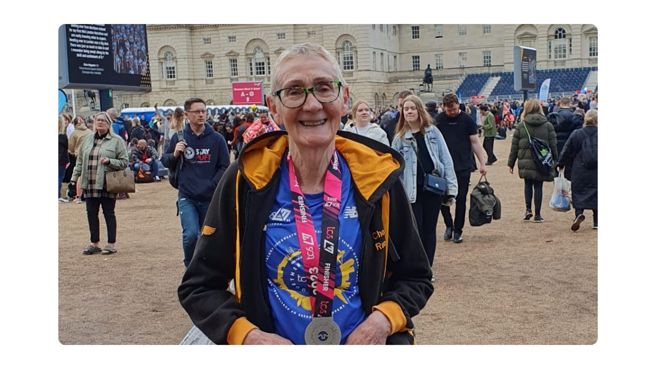 An older woman woman stands and smiles at the camera. She is wearing a medal and there are lots of people walking around behind her.