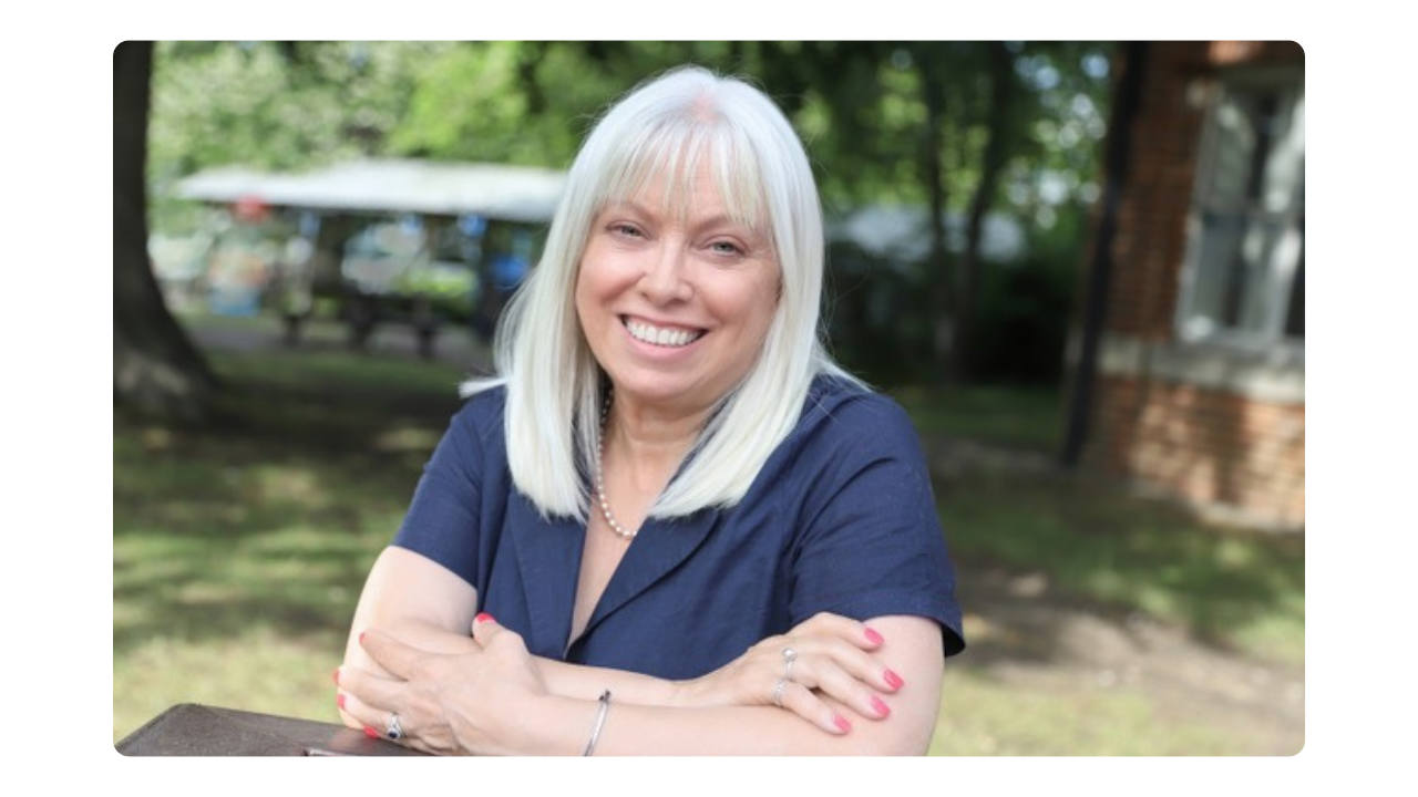 A woman sits outside and smiles at the camera with her arms crossed, leaning on a table. The background features sun, grass and trees.
