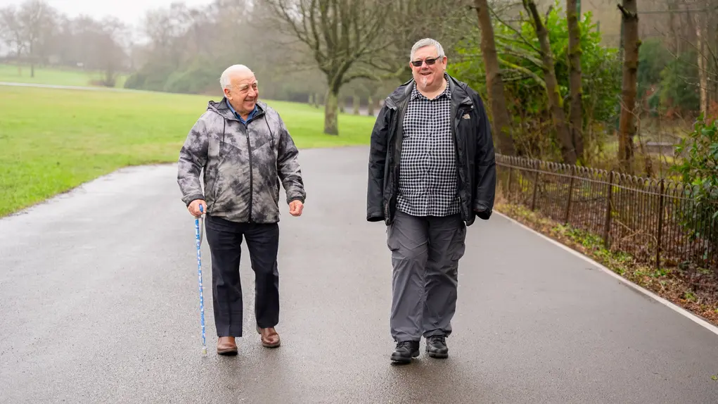 Two older men walking together along a path surrounded by grass and trees.