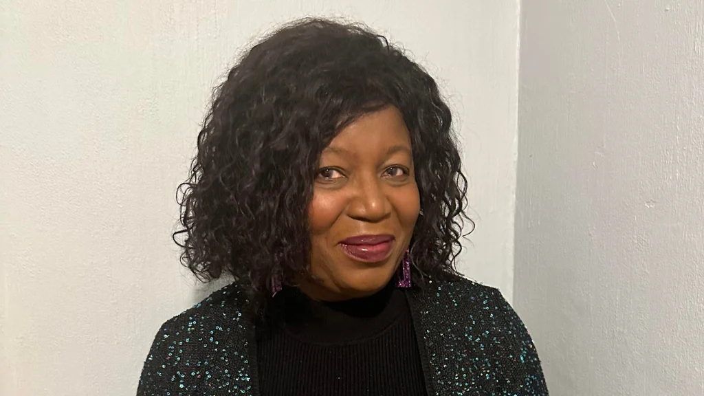 A woman stands in front of a white wall and smiles at the camera. She is wearing a sparkly black cardigan and black turtleneck.