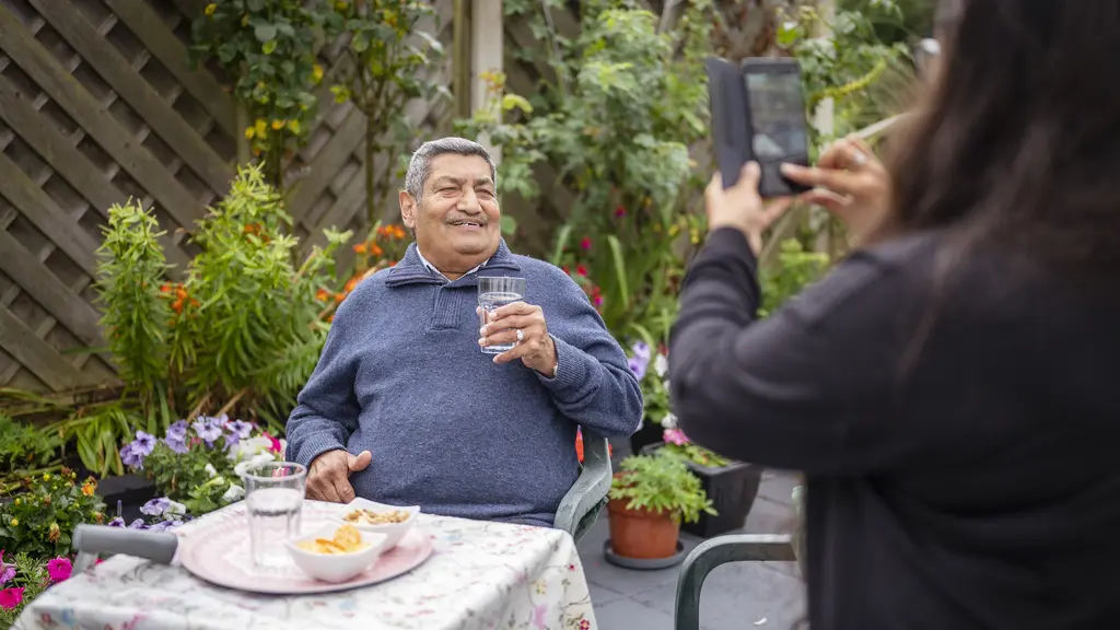 An older man sitting at a table outside smiling as someone takes his picture.