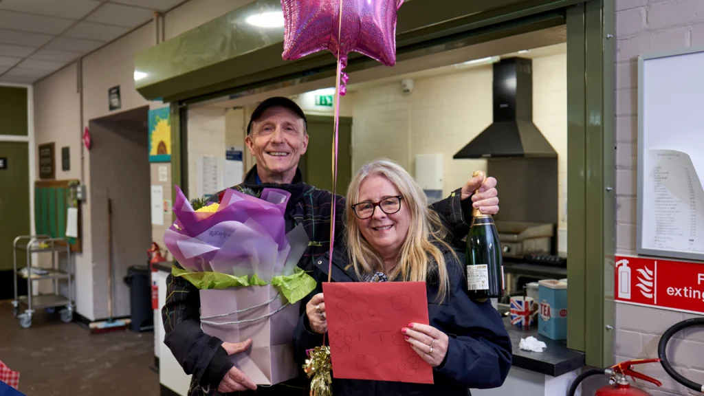 A man holds a star shaped balloon, flowers and prosecco and stands next to a woman holding a card. They are in a kitchen office environment.