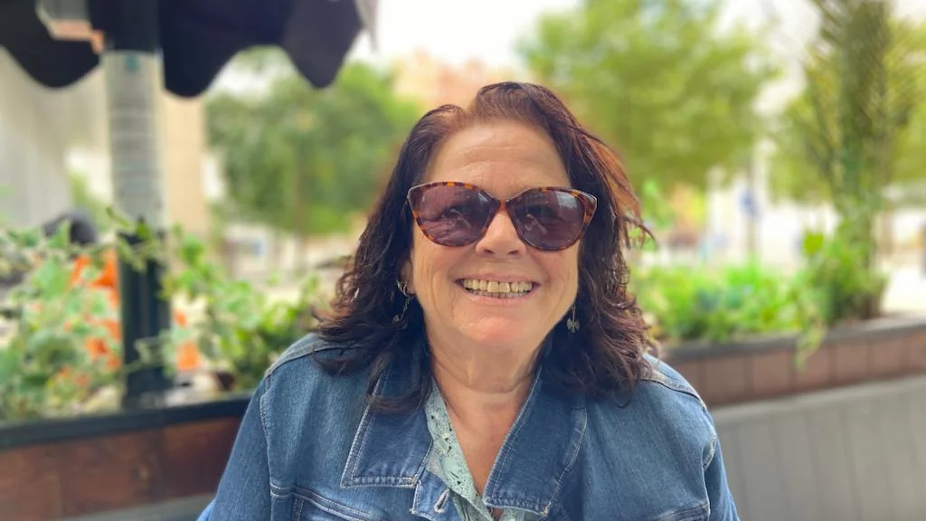 A woman wearing a denim jacket and sunglasses smiles while sitting outside.