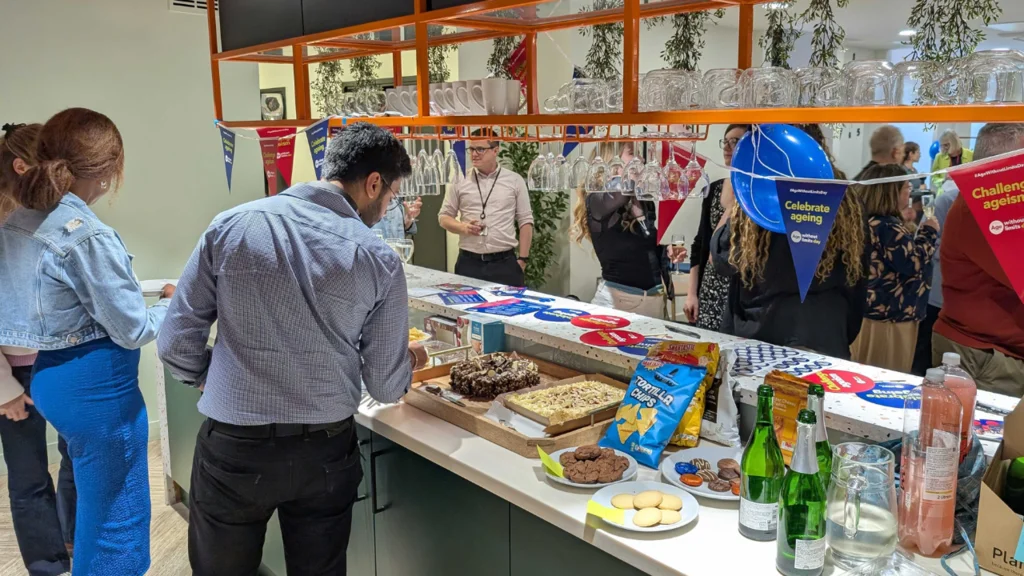A counter with food and drinks and Age Without Limits balloons and bunting, with people chatting in the background