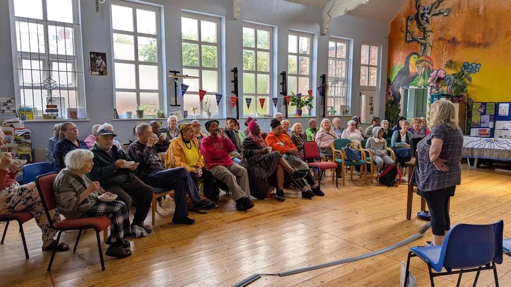 an older woman speaks to a crowd sat on chairs in a semi-circle