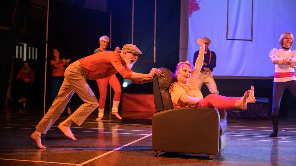 A group of older people perform a circus show, with a man pushing a woman in a chair across the stage