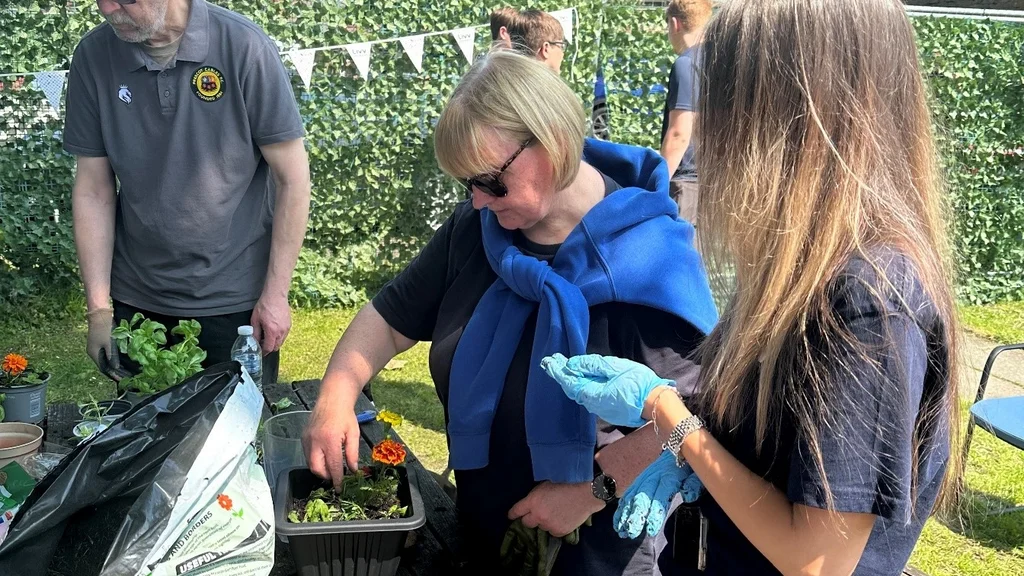 Older and younger people are potting plants at a garden table, which has trowels and gardening gloves on it