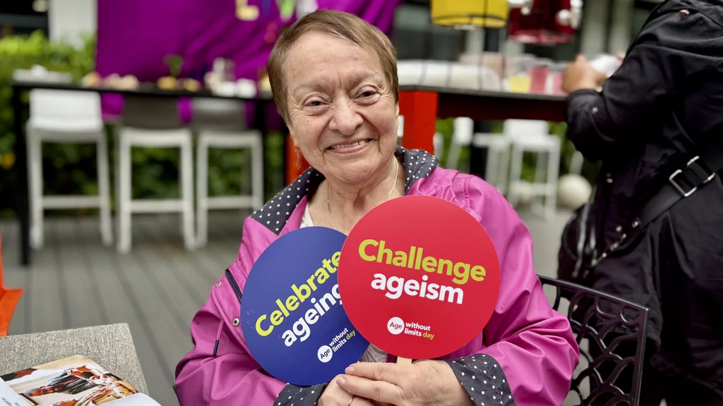 An older woman holds two Age Without Limits photo props which say celebrate ageing and challenge ageism