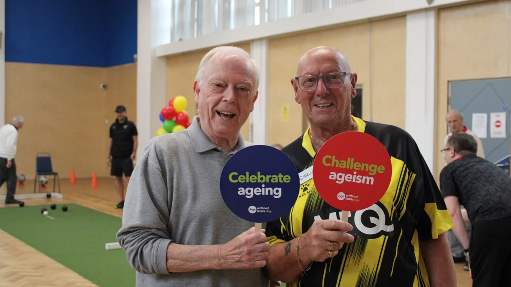 Two older men hold up Age Without Limits Day photo props saying celebrate ageing and challenge ageism. They are inside a gym hall with a sports activity behind them.
