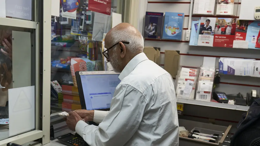 An older man with white hair handles cash behind a post office counter
