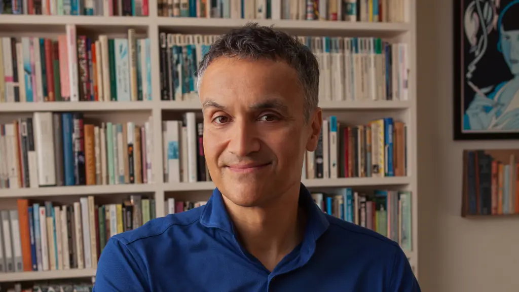 A man wearing a blue shirt smiles in the foreground with a bookshelf behind him
