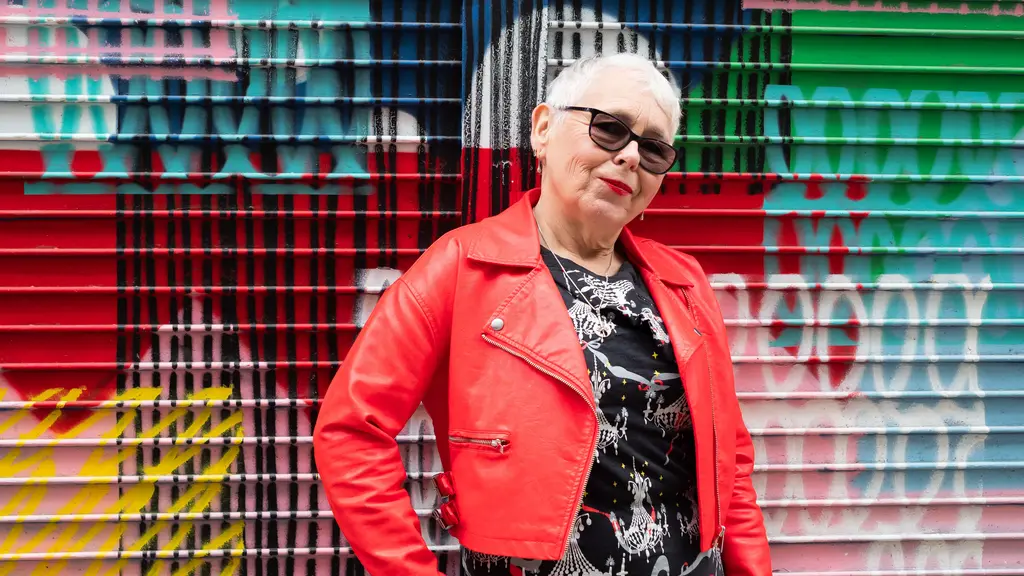 An older woman with short silver hair and a red leather jacket and sunglasses stands in front of graffiti art with her hand on her hip, looking at the camera.
