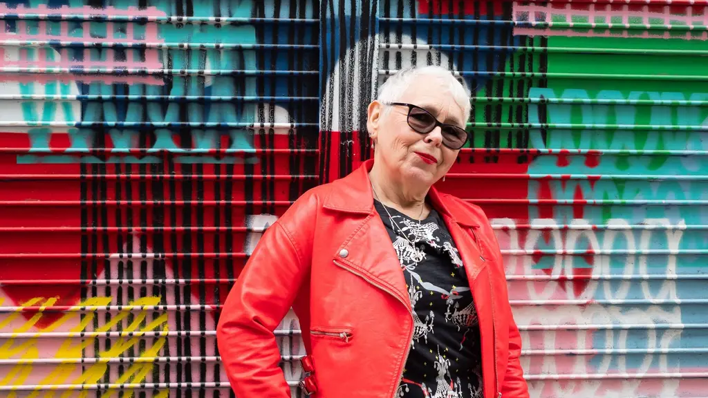 An older woman with short silver hair and a red leather jacket and sunglasses stands in front of graffiti art with her hand on her hip, looking at the camera.