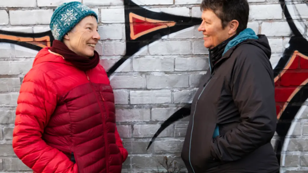 Two older women in winter coats lean against a wall with graffiti