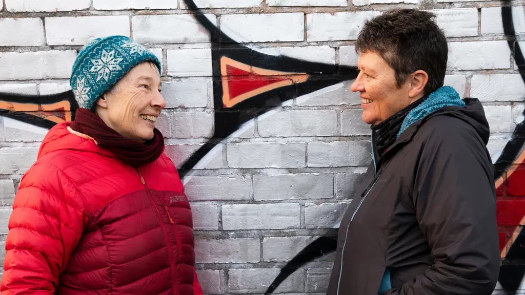 Two older women in winter coats lean against a wall with graffiti
