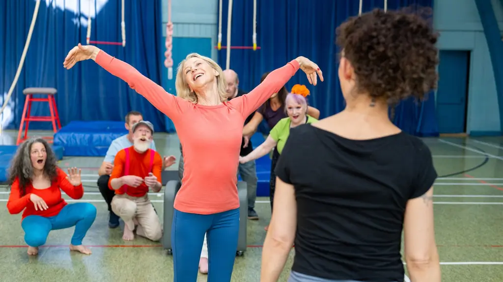 A woman wearing exercise clothes stands with her arms above her head as she rehearses for a circus show, while a group of people act shocked behind her.