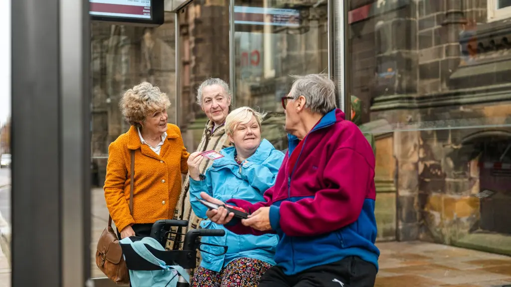 Four older people are standing and sitting at a bus stop. One man is talking and three women are listening.