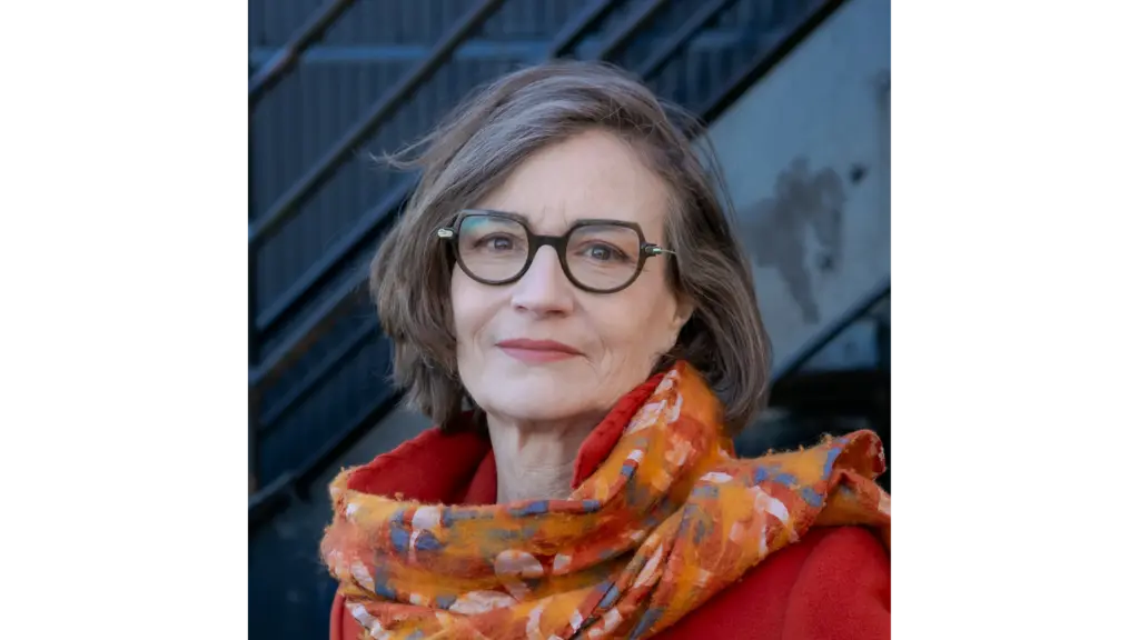 A woman with glasses, a bright orange coat and patterned orange scarf looks at the camera. A metal staircase is behind her.
