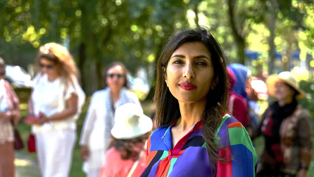 A woman in a bright shirt in a park looks directly into the camera, with other women blurred in the background