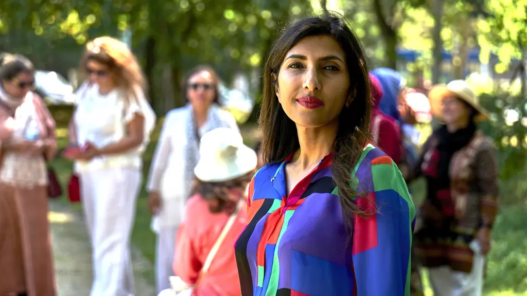 A woman in a bright shirt in a park looks directly into the camera, with other women blurred in the background