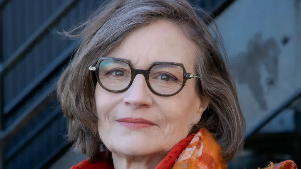 A woman with glasses, a bright orange coat and patterned orange scarf looks at the camera. A metal staircase is behind her.
