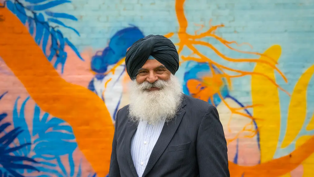 An older man wearing a turban and a suit stands in front of a colourful mural on a wall.