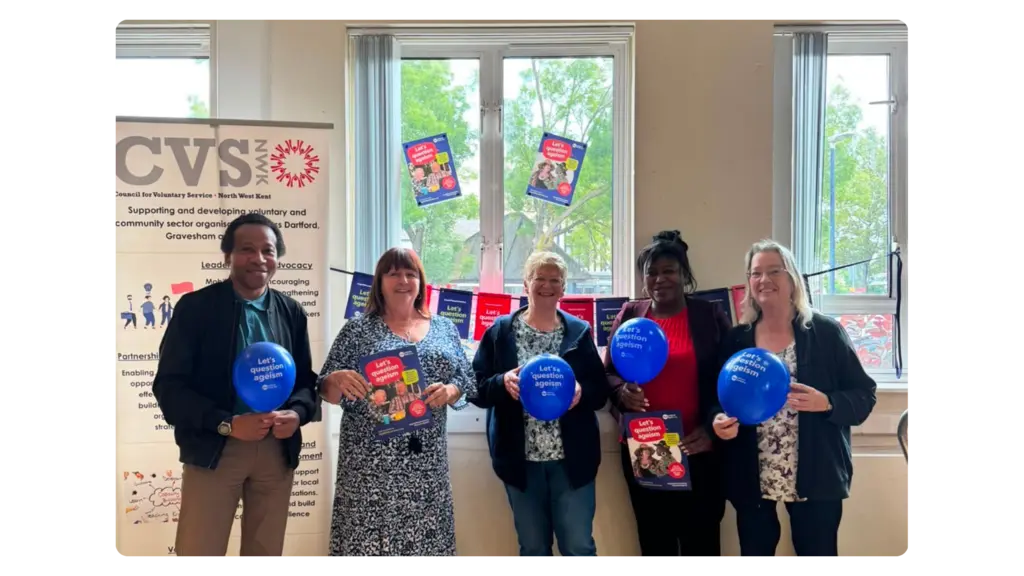 A group of five people stand in front of Age Without Limits Day bunting and hold Age Without Limits Day bright blue balloons