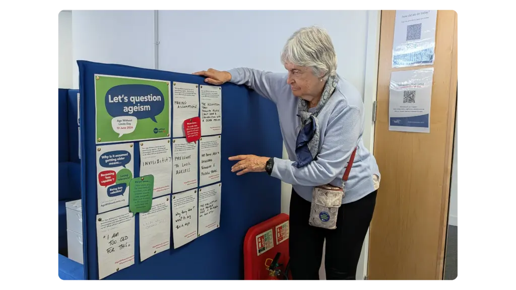 A woman leans on and points at a noticeboard full of Age Without Limits materials and people's reflections about age and ageism
