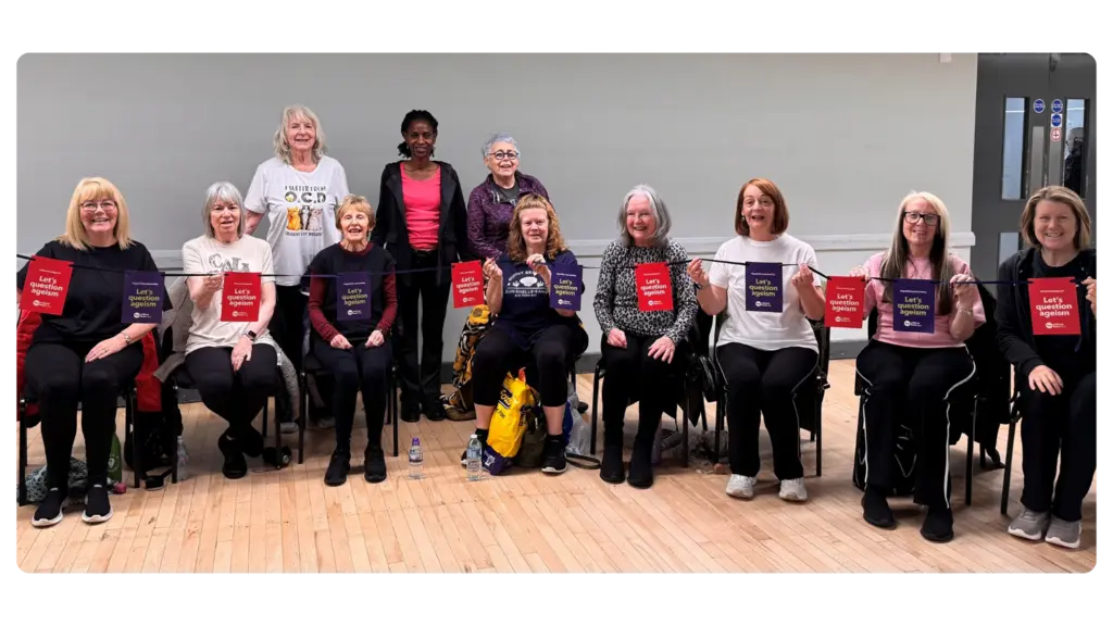 A group of women sit in a line on chairs and smile at the camera while holding up Age Without Limits bunting