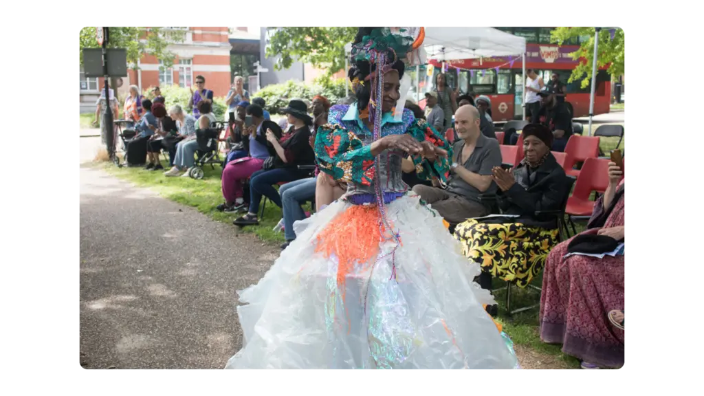 A woman walks down a catwalk in a park with a crowd in the background. she is wearing a brightly coloured dress made of recycled materials