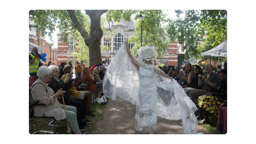 A woman holds out her arms in a flowing white dress and walks down a catwalk in a park with a crowd in the background