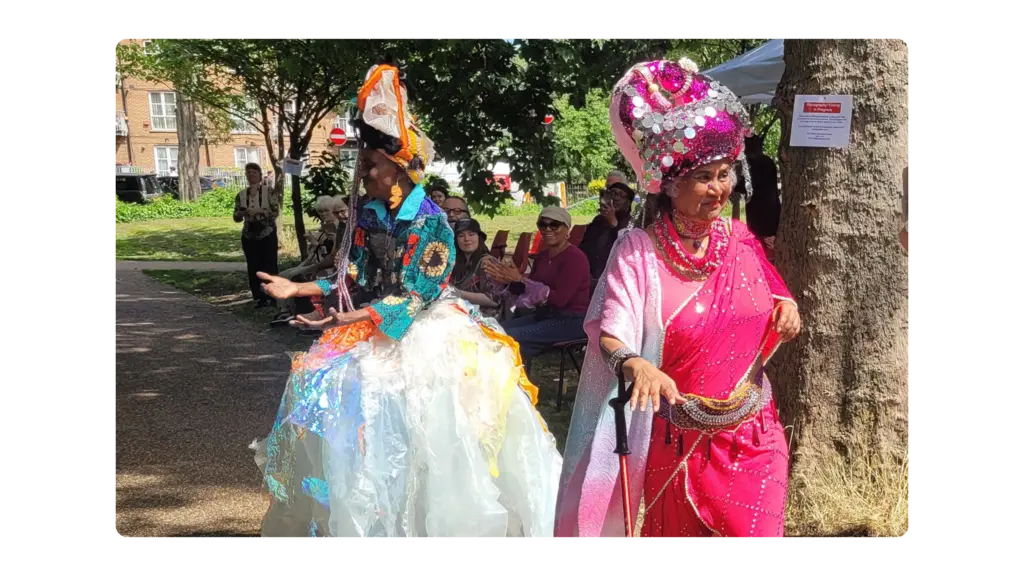 Two older women in brightly coloured clothes walk down a catwalk in a park with a crowd in the background