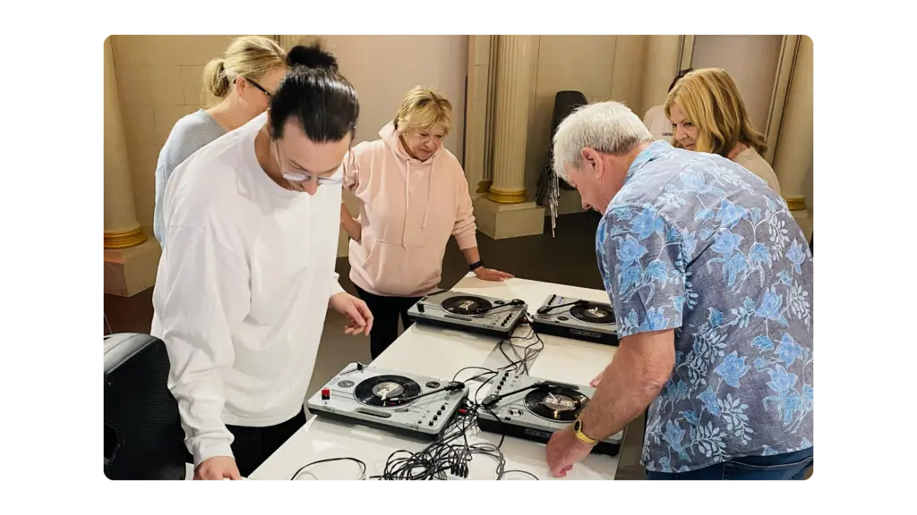 An intergenerational group of people stand in front of CDJs and learn how to DJ