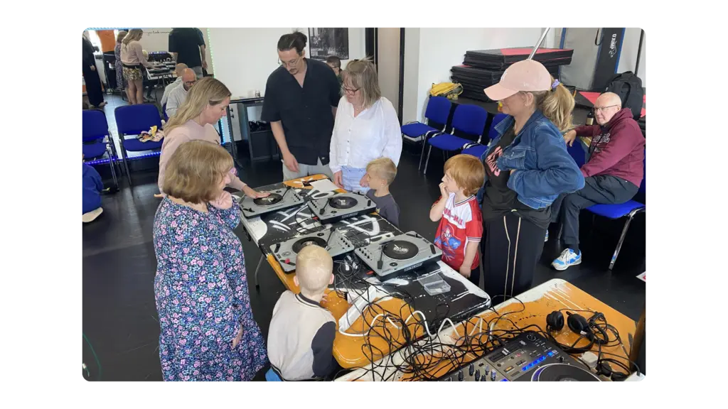 An intergenerational group of people stand in front of CDJs and learn how to DJ