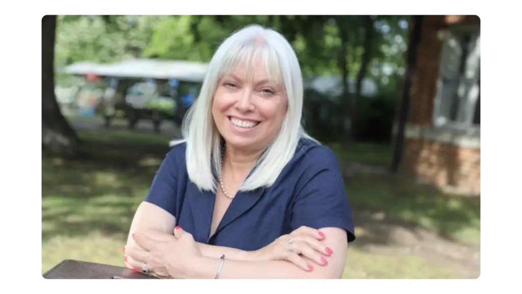 A woman sits outside and smiles at the camera with her arms crossed, leaning on a table. The background features sun, grass and trees.