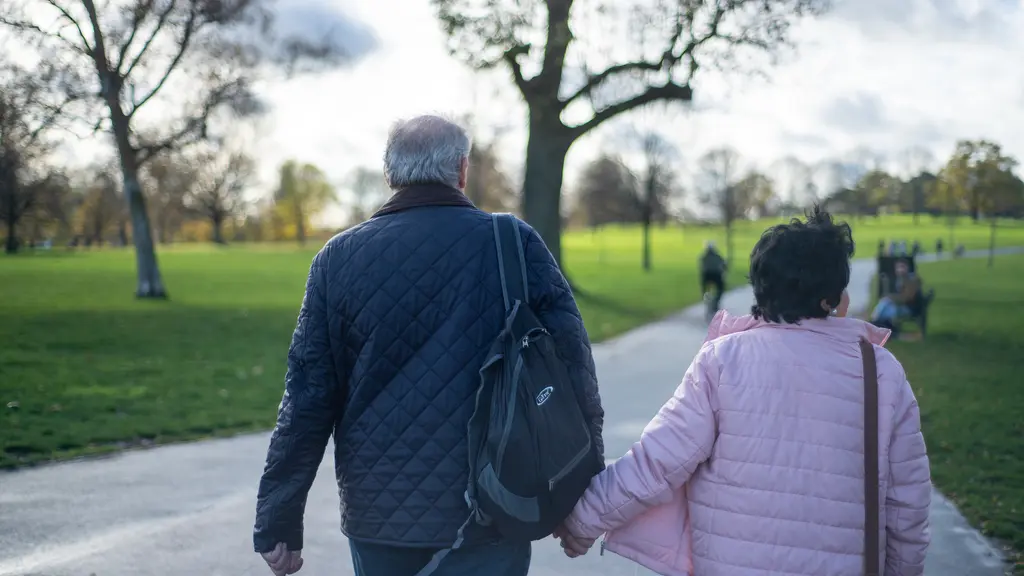 An older couple walking through a park, holding hands. The image is taken from behind.