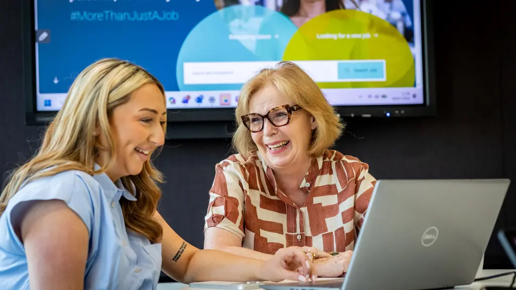 An older woman and younger woman work together at a laptop, with a presentation screen behind them. They are both smiling and laughing.