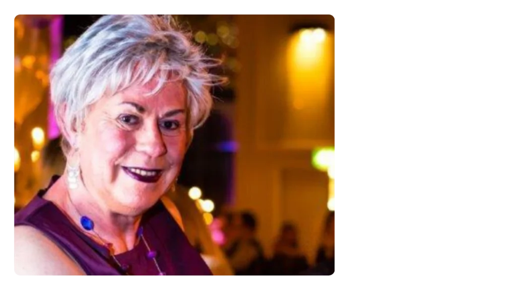 A woman wearing a purple dress smiles at the camera, with soft lighting and people behind her.