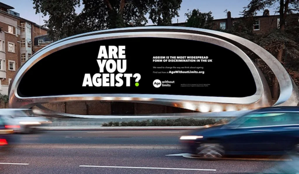 A roadside billboard saying Are You Ageist with cars driving past in front of it